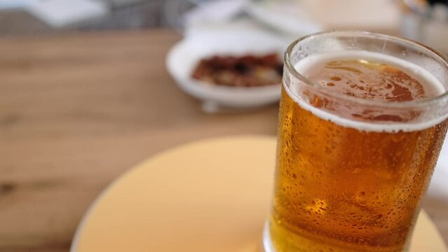 Close-up glass of cold beer on the table with snacks. Glass full light craft beer spin with drop of condensation cold drink. Cold drink glass on table. Cold alcohol stout beer drink in mug.