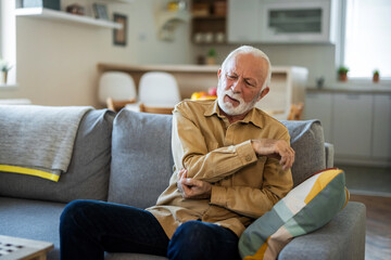 Unhappy senior man suffering from elbow pain at home. People, health care and problem concept. Senior man with arm pain.