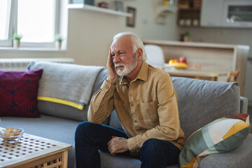 Depressed senior man sitting in the living room. Frustrated senior man with headache covering face and contemplating.