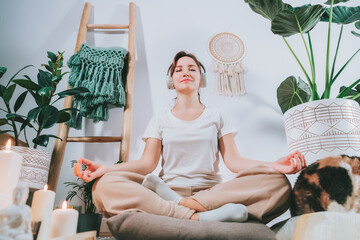 Young healthy woman practicing meditation at home, wearing headset, sitting in lotus position on...