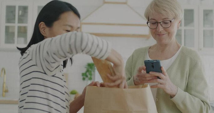Asian woman unpacking fresh healthy food from paper bag while senior lady smiling talking and using smartphone in kitchen at home. Groceries delivery and helping elderly people concept.