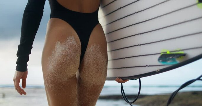 Young Caucasian female surfer walks on beach outdoors with surfboard. Woman in black bikini walks to ocean on beautiful sea water. Concept of recreation, tourism, healthy lifestyle, physical exercise.