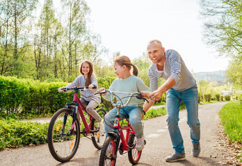 Obraz premium Smiling father with two daughters during outdoor walk. He teaching younger girl to ride a bicycle. They enjoy togetherness in the summer city park. Happy childhood concept image.