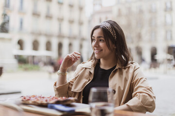 Portrait of happy beautiful young hispanic curvy woman sitting at restaurant. Smiling latin girl eating pizza. Pretty fashionable Spanish lady having dinner at cafe outdoors. - obrazy, fototapety, plakaty