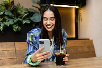 Happy zoomer multiethnic teen girl checking social media holding smartphone at cafe. Smiling young ...