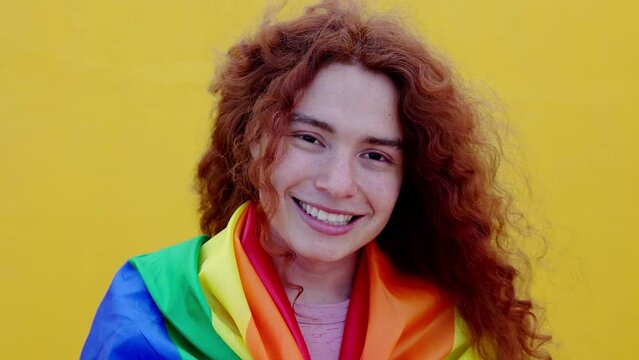 Portrait of red haired non binary man with rainbow flag smiling at camera over yellow background. LGBT community, diversity, transgender and non binary people concept
