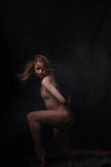 Ballerina in a beige bodysuit. Dark background. Sculpted beautiful female body. Pose of a gymnast.