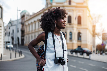 Young black female tourist enjoys walking through the streets of a beautiful European city. She is happy and using her photo camera to take fantastic architecture photographs. Bright sunny day.