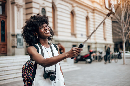 Young black female tourist enjoys walking through the streets of a beautiful European city. She is happy and using her smart phone to take selfie portraits and videos. Bright sunny day.