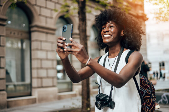 Young black female tourist enjoys walking through the streets of a beautiful European city. She is happy and using her smart phone to take some photos and videos. Bright sunny day.