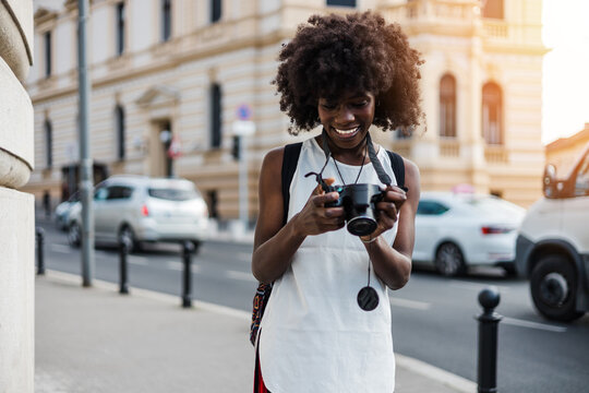 Young black female tourist enjoys walking through the streets of a beautiful European city. She is happy and using her photo camera to take fantastic architecture photographs. Bright sunny day.