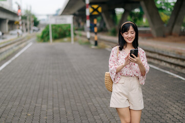 Naklejka premium Asian young woman traveler with weaving basket using a mobile phone beside railway train station in Bangkok. Journey trip lifestyle, world travel explorer or Asia summer tourism concept.