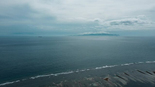 Aerial view of ocean shore against backdrop of seascape with a passing cargo ship and silhouettes of island Lombok with blue sky. Landscape of the tropical coast at low tide. Indian ocean trade routes