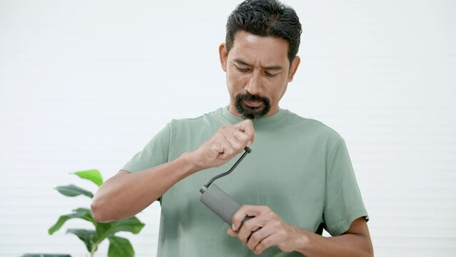 Asian man is standing grinding coffee beans with small grinder, which can be done by turning in circular motion until fineness is desired, on holiday morning, in order make coffee drink in morning.