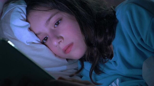 Teen girl scrolling social media texting on smartphone in bed in bedroom at night. Technology, internet, communication and people concept. Dependence on social networks in children. Not healthy sleep.
