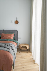 Bedroom interior with rust color linen and cushions on a bed, grey blanket, rattan bedside table and metal wall light.