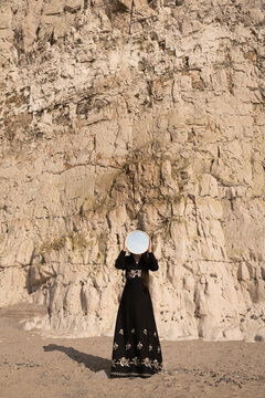 art portrait of girl in classic black dress near rocky wall holding mirror 