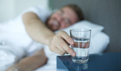 Young handsome man in the bed in bedroom at home in the morning lying under white blanket and drinking pure fresh water from glass 