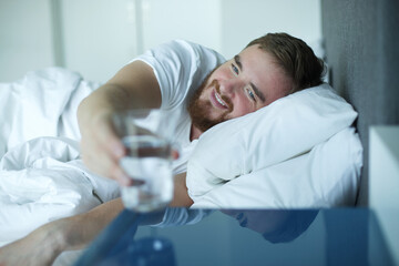Obraz premium Young handsome man in the bed in bedroom at home in the morning and drinking pure fresh water from glass 