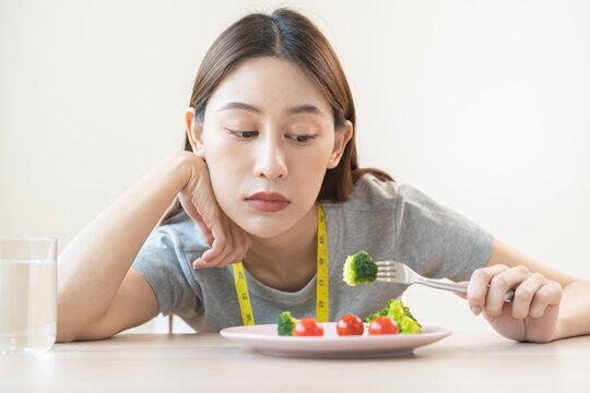 Anorexia, unhappy beautiful asian young woman, girl on dieting, hand holding fork at broccoli in salad plate, dislike or tired with eat fresh vegetables. Nutrition of clean, healthy food good taste.