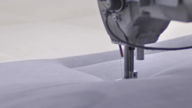 Worker sewing edges of mattress with help equipment in factory indoors.Worker sews cloth on mattress in factory indoors. keeps by hands white material while using equipment to stitch edges of product.
