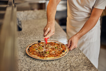 Close-up of a male worker, cutting a delicious pizza, using a pizza knife.