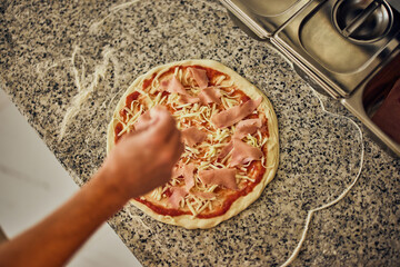 Top view of a male hand, putting a cheese on the pizza dough, making pizza for customers.