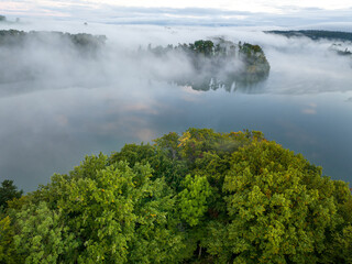 Aerial view of fog above lake early in the morning during sunrise. Misty scenery with reflections.