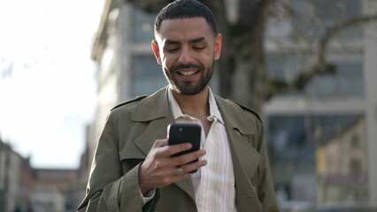 One happy Moroccan man smiling at social media content online while holding smartphone device standing in city street. Closeup face of a Middle Eastern man in tracking shot with motion