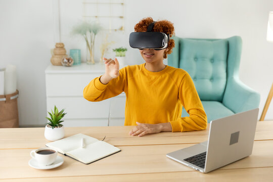 African american young woman wearing using virtual reality metaverse VR glasses headset at home. Girl touching air during VR experience on virtual reality helmet. Simulation hi-tech videogame concept
