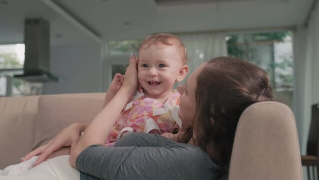 Adorable baby girl in smart dress lying on her mother chest and laughing while both having fun and spending leisure together in home environment