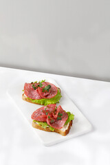 Sandwich, toast with sliced salami, sausage, green lettuce on white cutting board. Hard light, morning sunny breakfast. Snack, bruschetta.