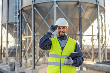 A industry supervisor is holding tablet in his hands and greeting while standing in front of the silos full of supplies.