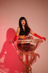 Pinup girl standing near big glass. Black wig, black underwear, stockings and red gloves, red and blue light, red lipstick. Beautiful posch woman sitting near huge plastic goblet.