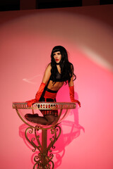 Pinup girl standing near big glass. Black wig, black underwear, stockings and red gloves, red and blue light, red lipstick. Beautiful posch woman sitting near huge plastic goblet.