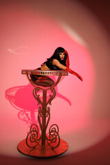 Pinup girl standing near big glass. Black wig, black underwear, stockings and red gloves, red and blue light, red lipstick. Beautiful posch woman sitting near huge plastic goblet.