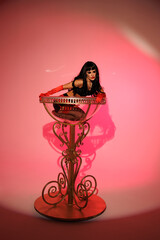 Pinup girl standing near big glass. Black wig, black underwear, stockings and red gloves, red and blue light, red lipstick. Beautiful posch woman sitting near huge plastic goblet.