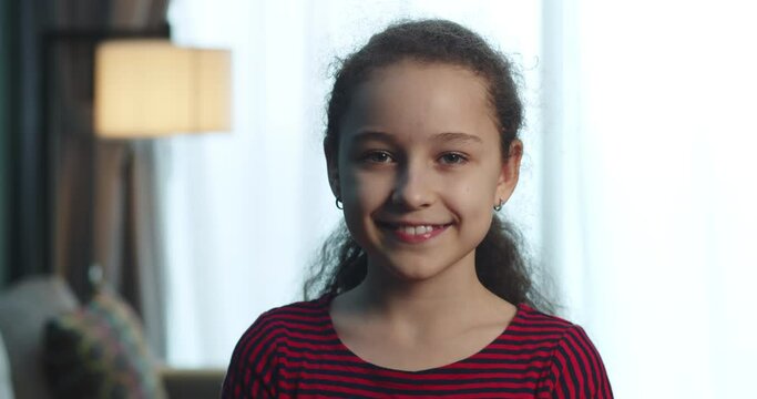 Portrait of a beautiful little girl, a smiling child in a red dress, looking at the camera, sits on the sofa at home, a cute child with a beautiful face sits on the sofa. Happy childhood concept. 4K
