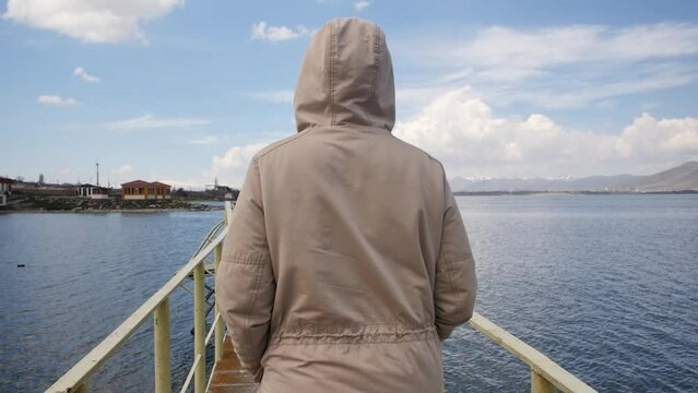 A young woman in a warm jacket, wearing a hood, walks along the sea pier looking at the sea and the mountain coast. A look behind a woman walking along the pier. Scenic mountain landscape and sea