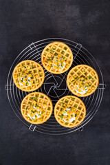 Small Round corn waffles with soft cottage cheese and greens. On a serving metal stand. Dark gray background. Top view