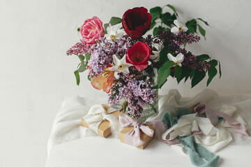 Stylish bouquet with gift box on rustic wooden table. Happy Mothers day. Spring lilac, tulips, daffodils arrangement with present on soft fabric in modern rustic room. Happy Womens day