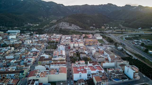 Spanish town surrounded by mountains. Features houses, apartments, and a highway, with warm sun and shadows.