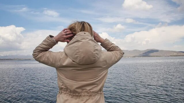 Caucasoid adult happy blonde enjoys the sea, fresh clean air and mountain landscape standing on the pier of the seashore and takes off the hood from her head enjoying life