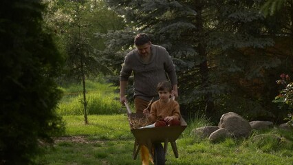 father rides son in a wheelbarrow with hay in the garden in spring time, having fun together