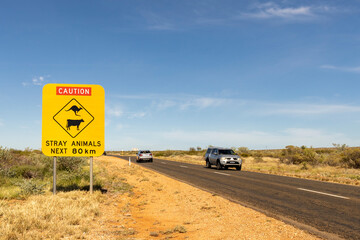 street sign "caution" and " stray animals next 80 km" with a kangaroo and cattle on it, cars driving in both directions on the road beside, travel australia