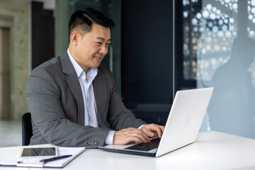 Successful mature asian working inside office using laptop, man typing on keyboard and smiling, businessman satisfied with work and achievement results, programmer at work.