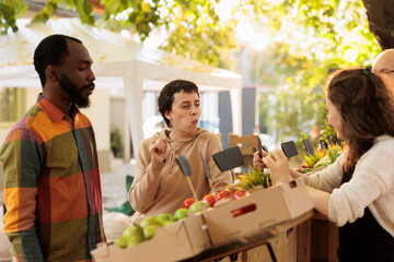 Young customers tasting free food samples at farmers market, fresh juicy fruits and vegetables outside. Man and woman buying healthy products from local fresh organic marketplace.