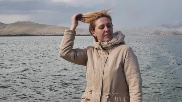 Portrait of a young adult happy woman in a warm jacket standing on the seashore with mountain shores and touching her hair on her head with her hand enjoying the wind and fresh sea.