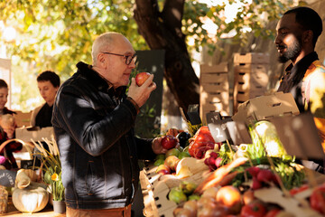 Elderly client smelling bio apples and standing near farmers food market stand with variety of fresh pesticide free fruits and vegetables. Old man buying locally grown organic produce. - obrazy, fototapety, plakaty