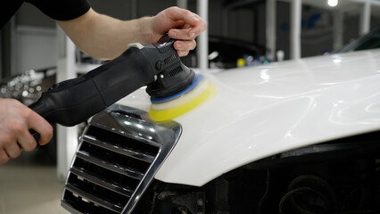 Hands of professional car service male worker, with orbital polisher, polishing white luxury car hood in auto repair shop. Car detailing and polishing concept.